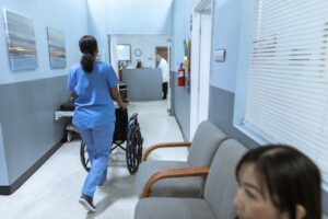 A nurse in scrubs pushes a wheelchair through a well-lit hospital corridor, attending to patient care.